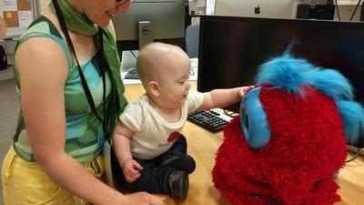 Jacqueline, a woman wearing glasses and a silky scarf, stands behind a baby seated on a table, looking down at the baby and smiling. The baby is reaching out to touch a fluffy Tega robot. In the background are monitors and office stuff.