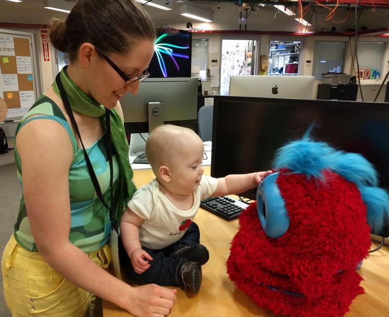 Jacqueline, a woman wearing glasses and a silky scarf, stands behind a baby seated on a table, looking down at the baby and smiling. The baby is reaching out to touch a fluffy Tega robot. In the background are monitors and office stuff.