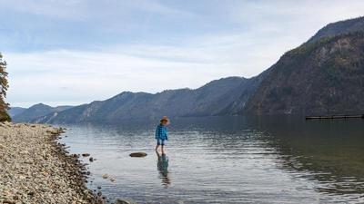 a young girl stands in a lake, pants rolled up to her knees, rocky shore behind her on the left, a mountain in the distance on the right