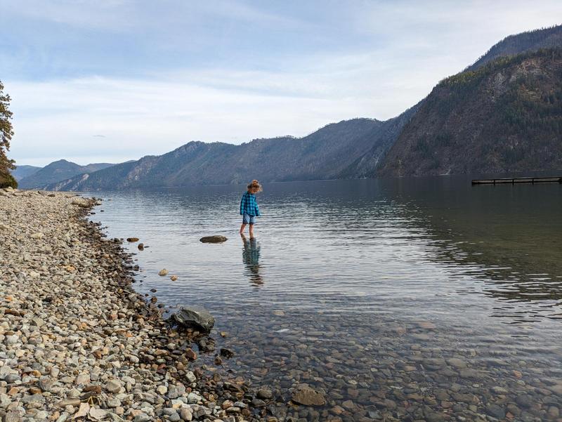 a young girl stands in a lake, pants rolled up to her knees, rocky shore behind her on the left, a mountain in the distance on the right