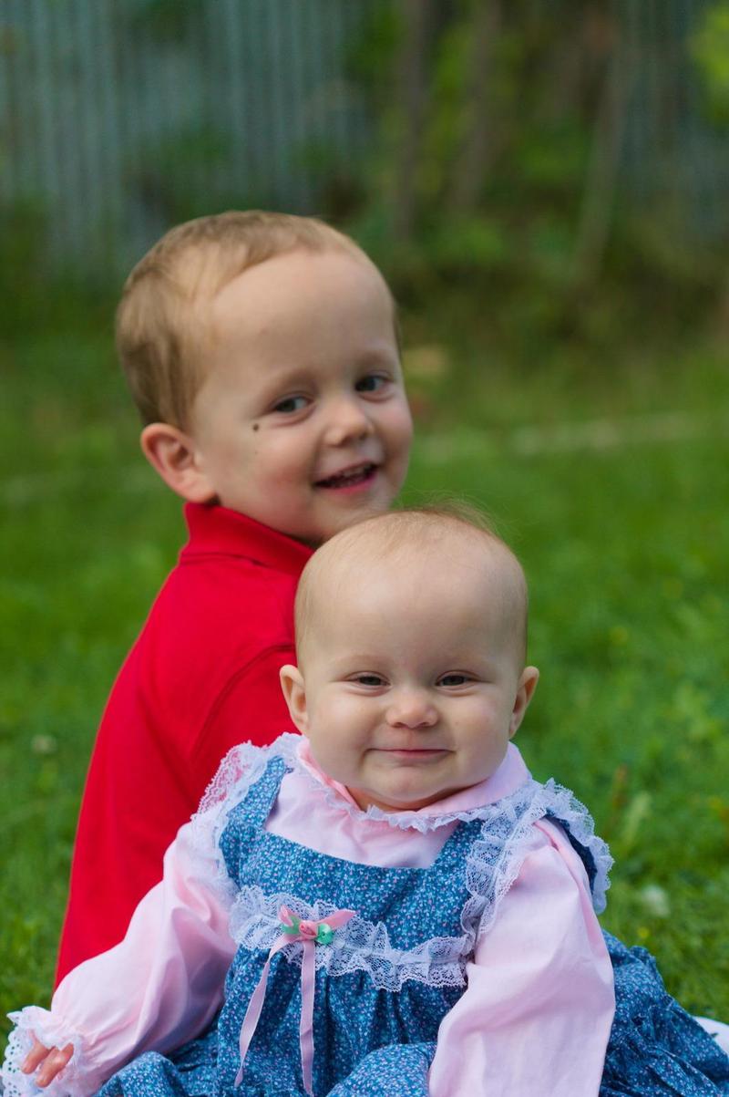 Three-year-old boy and six-month-old girl sitting in the grass together smiling