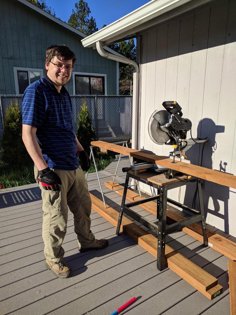 Randy standing on a deck in the sun next to a miter saw on a sawhorse loaded with a 2x6 plank of wood