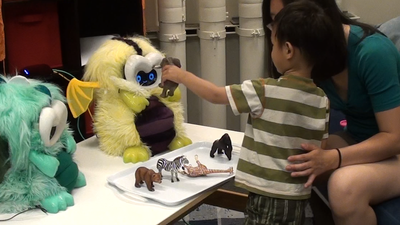 a young boy stands in front of his mom, who is seated and leaning forward, and holds up a toy elephant in front of a small robot's face