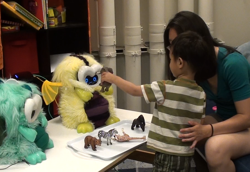 a young boy stands in front of his mom, who is seated and leaning forward, and holds up a toy elephant in front of a small robot's face