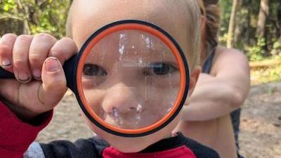 a child in a red shirt holds up a magnifying glass in front of his face, so his eyes and nose look really big