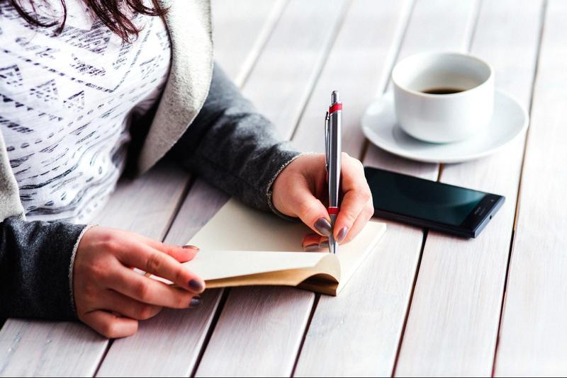 woman sitting at a table writing in a small journal with a cup of coffee