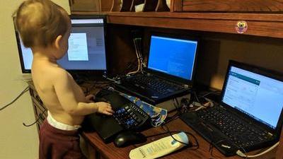 a toddler standing on a folding chair at a desk with three computers, pretending to type