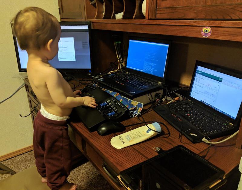 a toddler standing on a folding chair at a desk with three computers, pretending to type