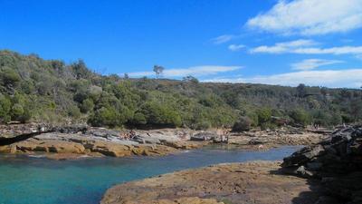 an inlet of bright blue water in sharp red-brown rocks at royal national park in sydney, australia, bordered by round green trees