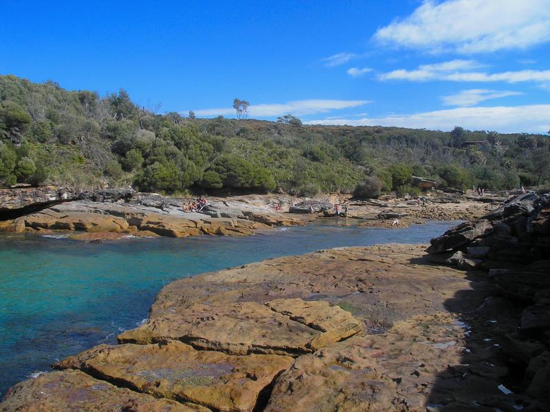 an inlet of bright blue water in sharp red-brown rocks at royal national park in sydney, australia, bordered by round green trees