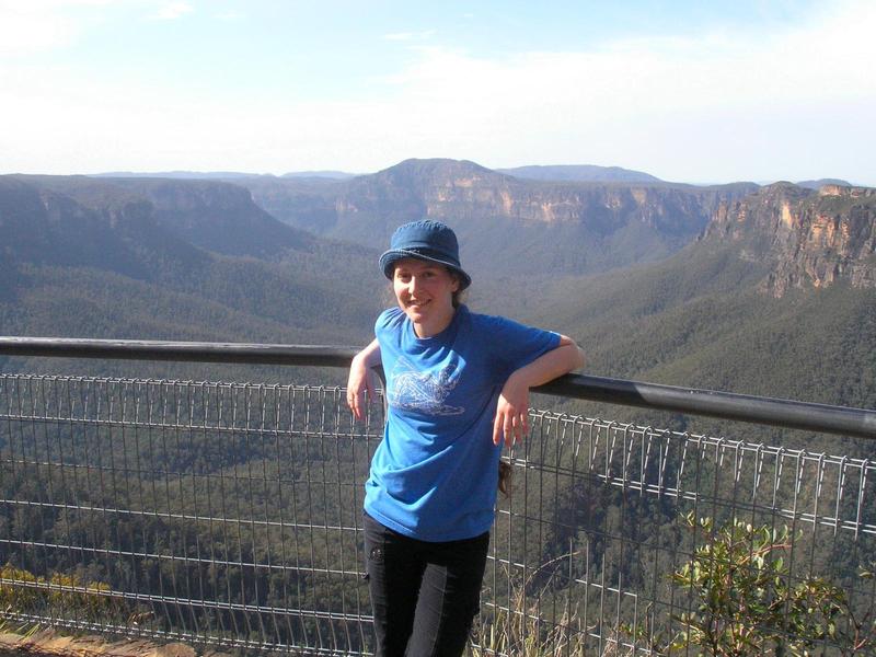 jacqueline leaning against a railing at a lookout in the Blue Mountains of Australia, tree-covered cliffs in the background