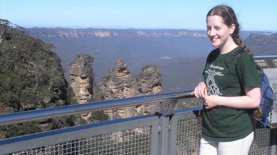 Jacqueline stands at a metal railing at a trailhead in the Blue Mountains, Australia, overlooking the steep hills and the three pillars of rock known as the Three Sisters