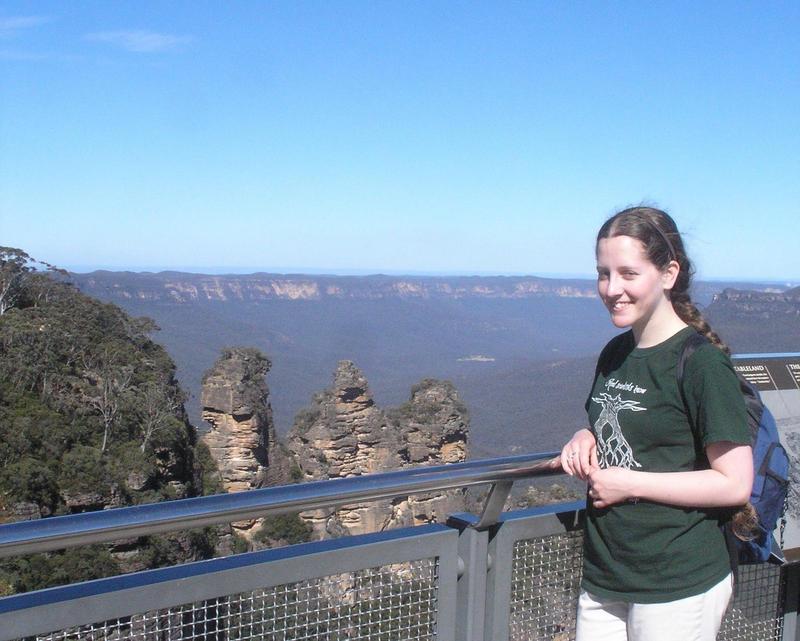 Jacqueline stands at a metal railing at a trailhead in the Blue Mountains, Australia, overlooking the steep hills and the three pillars of rock known as the Three Sisters