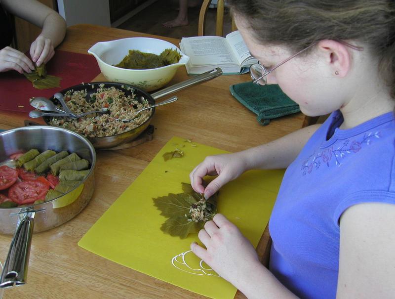 teenage jacqueline at a table folding a grape leaf around a blob of rice