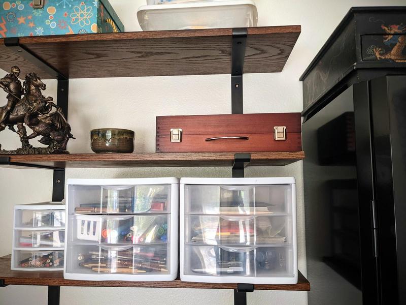 dark oak shelves on industrial brackets on a wall with boxes and drawers on them, beside a shiny black cabinet