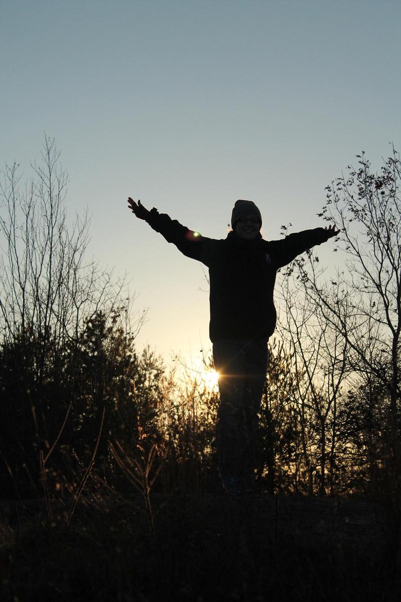 silhouette of a person with arms outstretched on wintery day, in front of bare-limbed trees at sunset