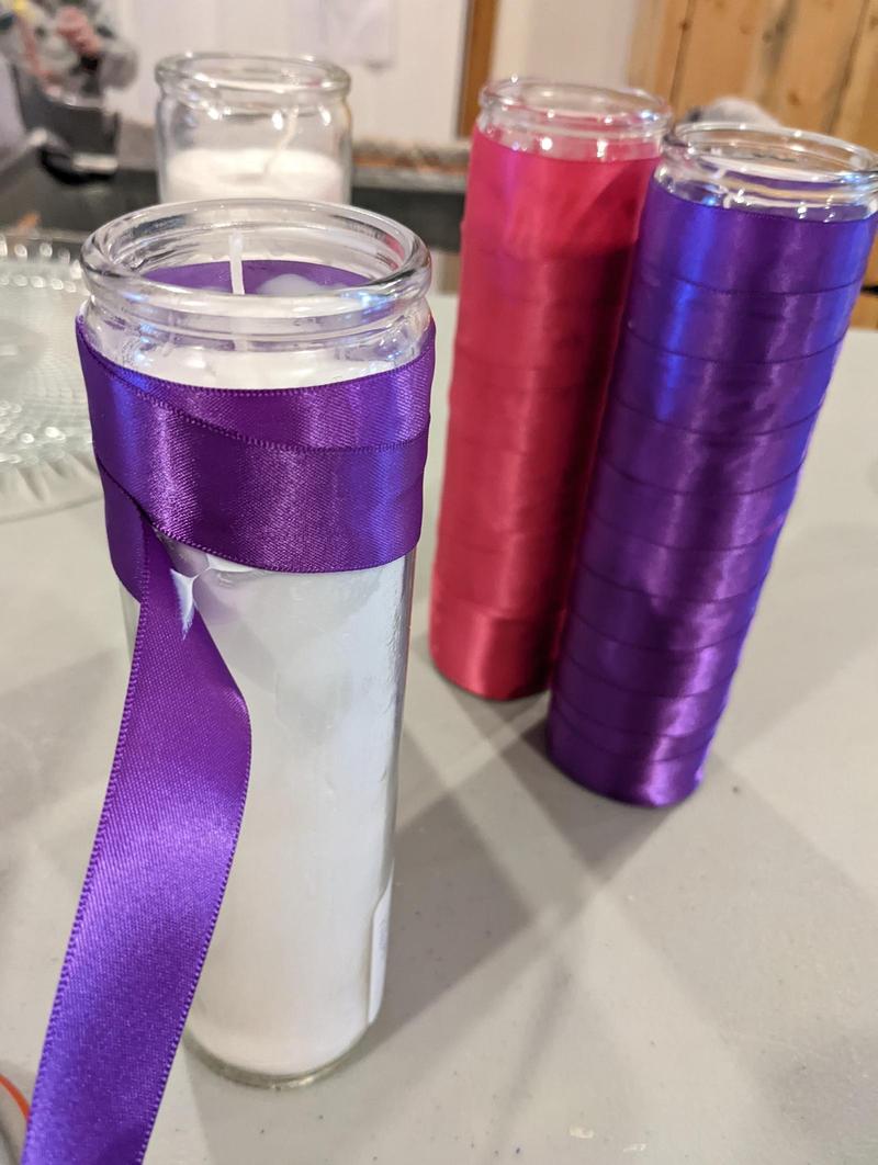 two tall candles wrapped in ribbon on a table with one candle only partially wrapped, with the ribbon still being glued