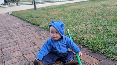 toddler sitting on a brick path by a lawn looking sideways, holding a large green pushbroom