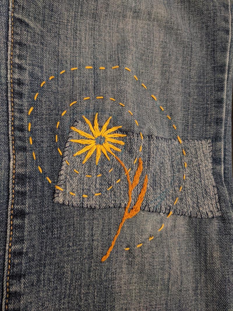 embroidered yellow flower with a brown stem and yellow dotted swirl around the flower over a rectangular patched knee of a pair of blue jeans