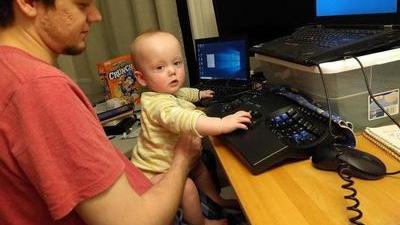Baby Elian stands on Randy's lap and types on his keyboard in front of three monitors
