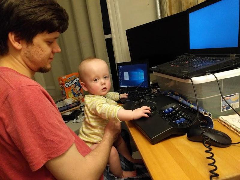 Baby Elian stands on Randy's lap and types on his keyboard in front of three monitors