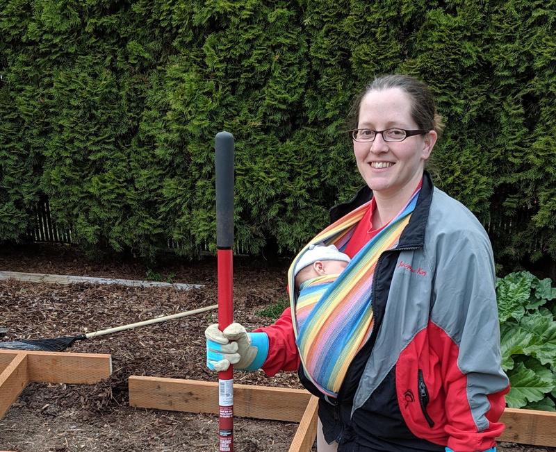 Jacqueline, wearing her baby in a woven rainbow wrap, stands in a backyard with a shovel and work gloves