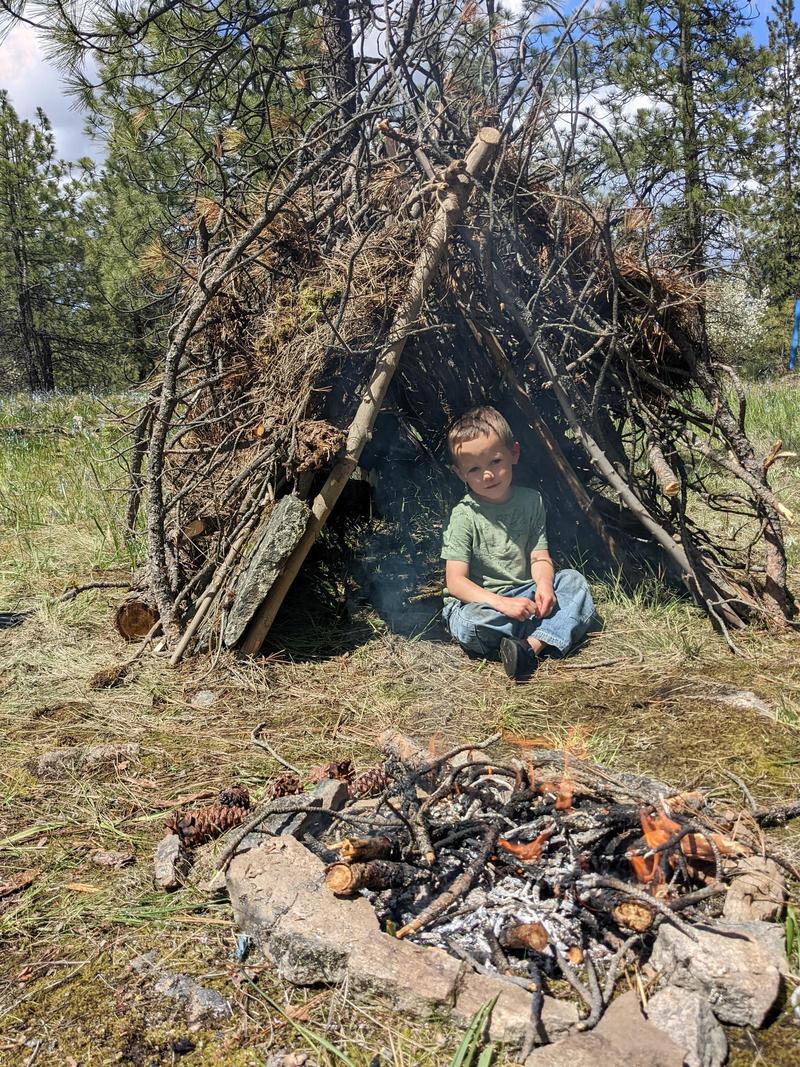 a five-year-old boy sits in the entrance to a triangular shelter made of branches, rocks, and moss, a small fire smoldering in a ring of rocks in the foreground