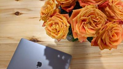 silver macbook rests on a wooden table beside a vase of bright summery roses