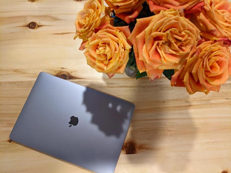 silver macbook rests on a wooden table beside a vase of bright summery roses