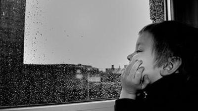 a young boy rests his chin on his hand in profile, gazing boredly out a rain-streaked window