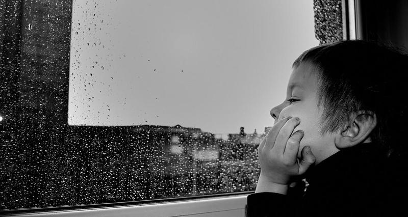 a young boy rests his chin on his hand in profile, gazing boredly out a rain-streaked window