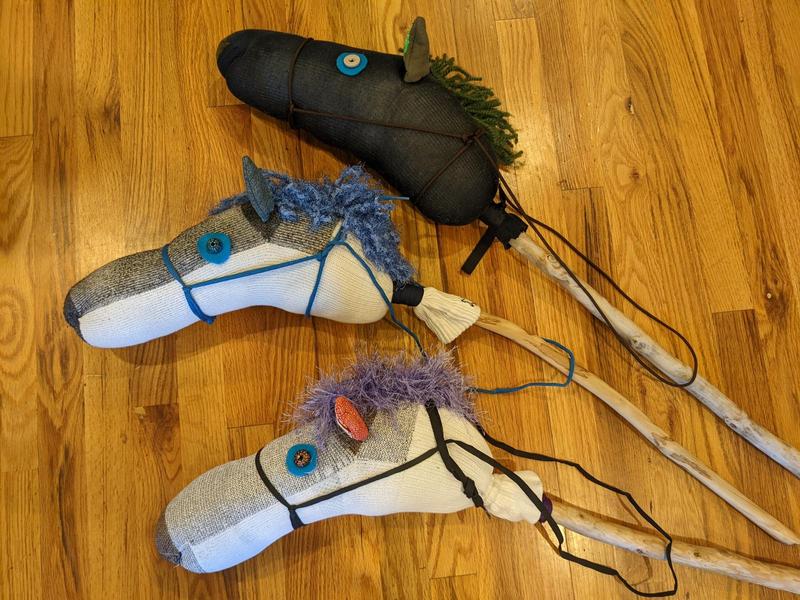 Three homemade sock hobby horses lined up on the floor, made with bright colors and fabrics that aren't particularly horselike