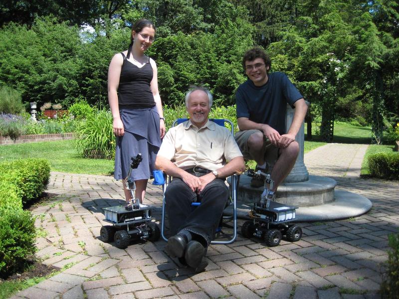 Jacqueline and Josh de Leeuw standing behind Ken Livingston, who sits in a chair, with two mechanical robots beside him