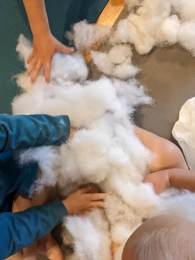 a pile of cotton stuffing in a baby's lap with hands from two other kids reaching to grab it or squish it