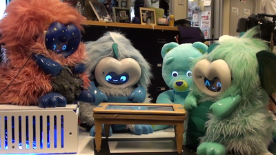 Four furry robots sitting around a wooden table inset with a tablet. From left to right, a red dragonbot, a blue dragonbot, a teddy bear robot, and a green dragonbot.