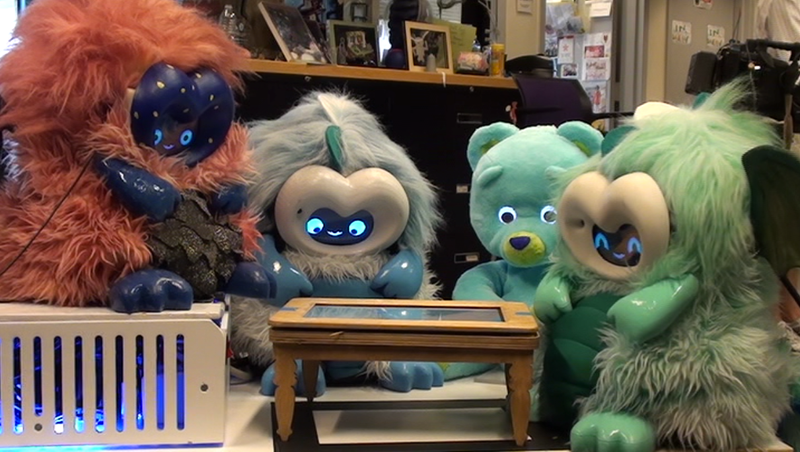 Four furry robots sitting around a wooden table inset with a tablet. From left to right, a red dragonbot, a blue dragonbot, a teddy bear robot, and a green dragonbot.