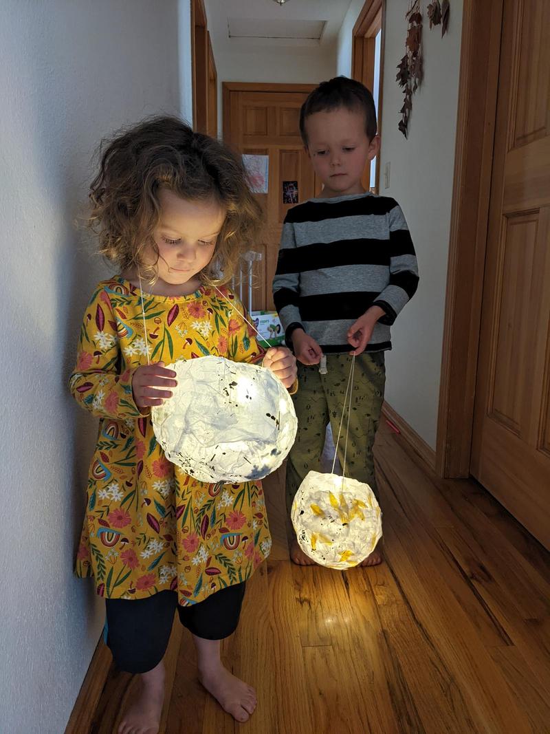 a three-year-old girl and five-year-old boy standing in a dim hallway holding glowing paper lanterns