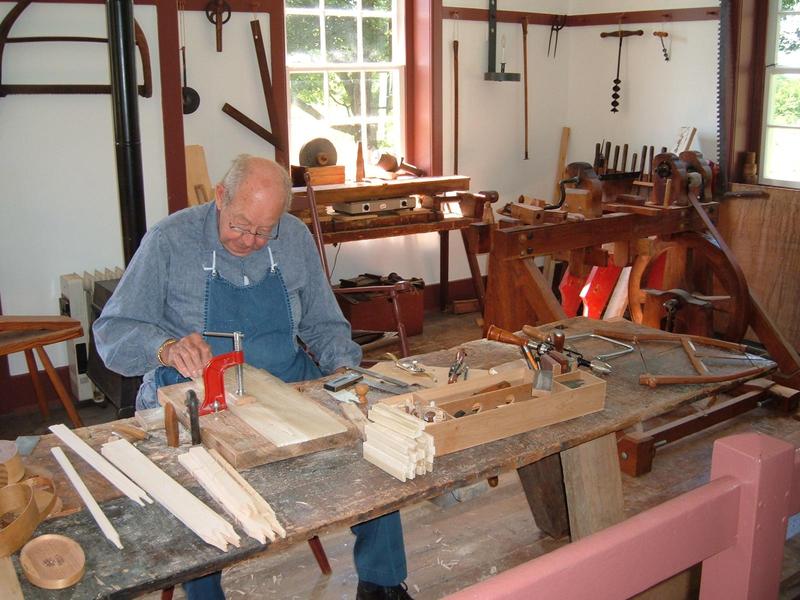 a mostly-bald older man sits at a workbench in a woodshop, clamping some pieces of wood together, old tools hanging on the wall in the background