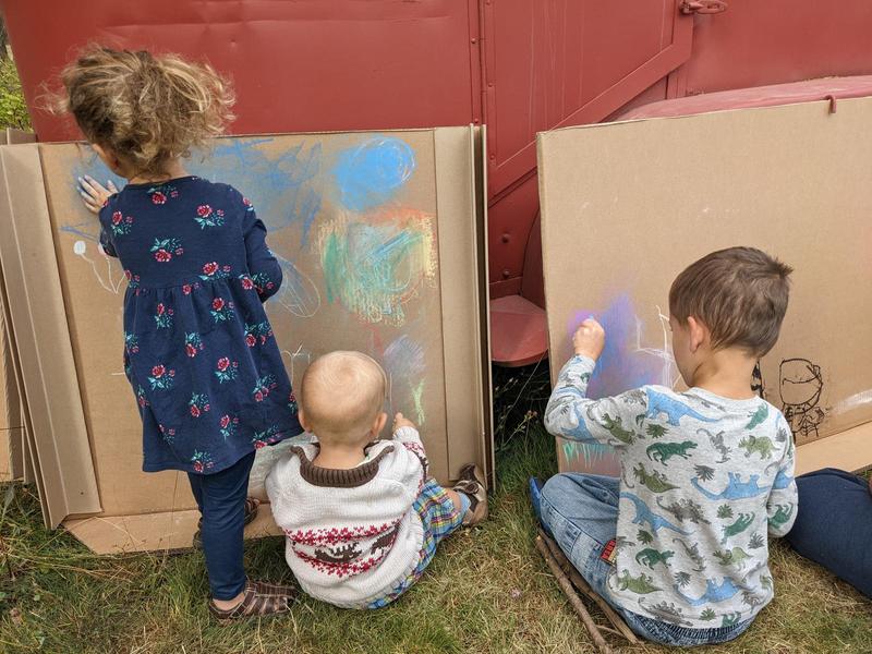 Three young children with their backs to the camera coloring with chalk pastels on large pieces of cardboard