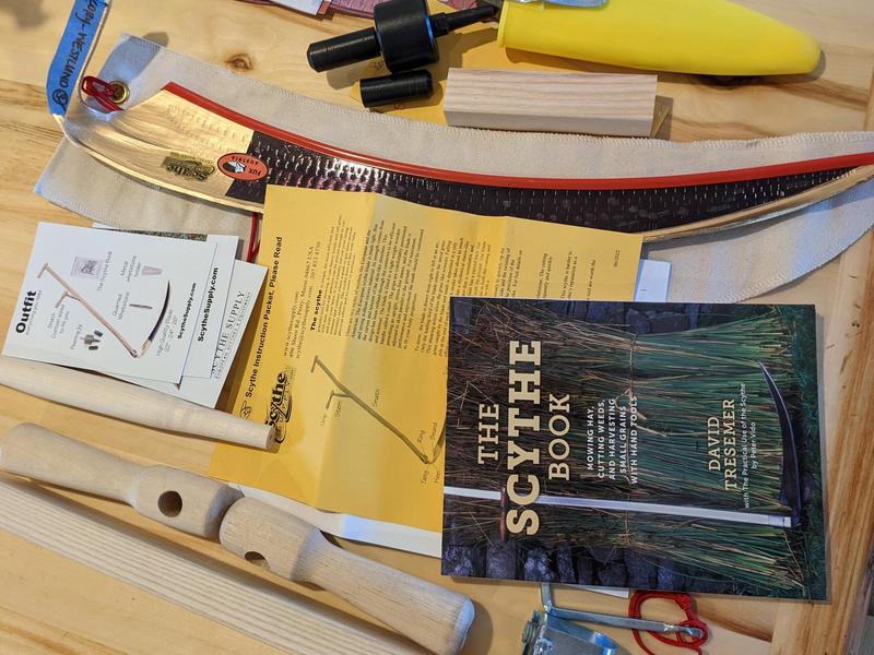 parts of a scythe laid on a table, alongside printed instructions, a book about scythes, and a peening jig