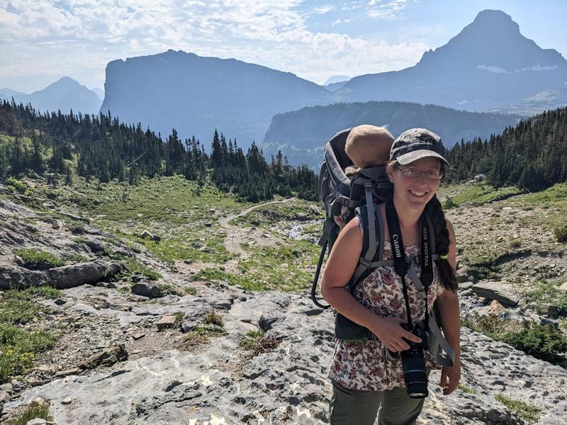 Jacqueline grinning and wearing a sleeping baby in a hiking backpack, framed by the pine forests and distant blue peaks of the Rocky Mountains in Glacier National Park