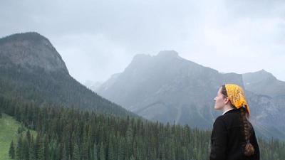 Jacqueline stands before a brilliant aqua blue alpine lake, with coniferous forests covering the slopes of mountains rising into cloudy mist on the opposite shore. She wears a black fleece and yellow banana; she looks up at the mountains, hands in pockets