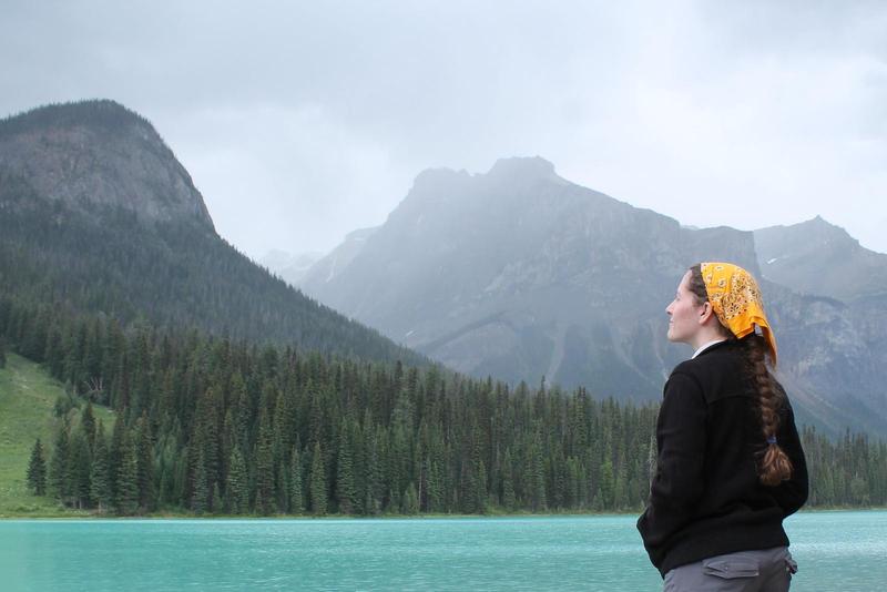 Jacqueline stands before a brilliant aqua blue alpine lake, with coniferous forests covering the slopes of mountains rising into cloudy mist on the opposite shore. She wears a black fleece and yellow banana; she looks up at the mountains, hands in pockets