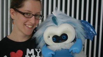 Jacqueline smiling, wearing glasses and a tshirt saying I <3 my, holding a blue dragonbot robot in front of a stripy background at the MIT Media Lab