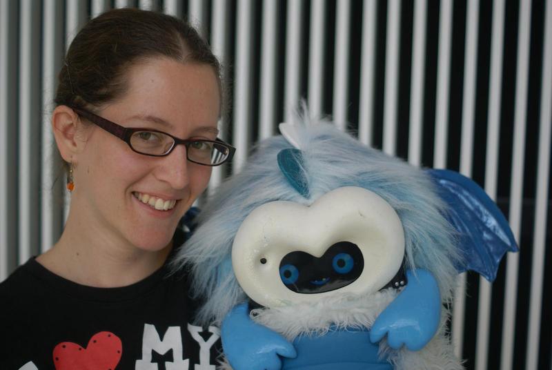 Jacqueline smiling, wearing glasses and a tshirt saying I <3 my, holding a blue dragonbot robot in front of a stripy background at the MIT Media Lab