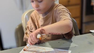 a three-year-old girl with her hair pulled back in a ponytail looks down intently as she draws on paper with an orange marker