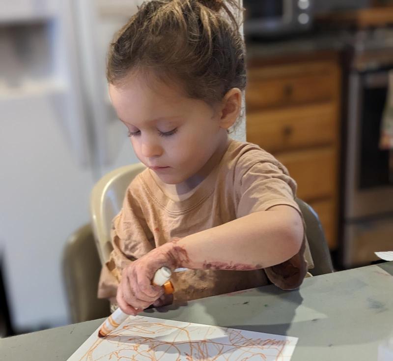 a three-year-old girl with her hair pulled back in a ponytail looks down intently as she draws on paper with an orange marker