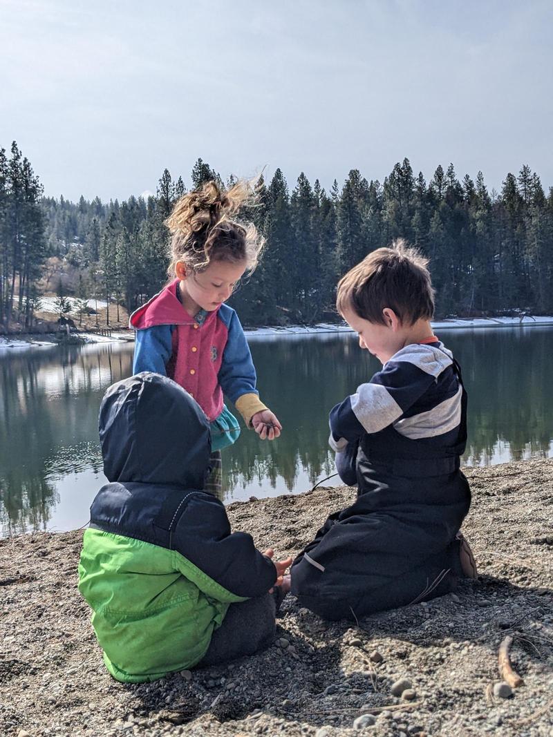 A little child in a coat sitting in the sand on a riverbank, looking toward a four-year-old girl with a ponytail who is standing and a six-year-old boy who is kneeling in the sand; the girl holds a handful of sand. In the background is a river and across the river is snow and pine trees on low hills.