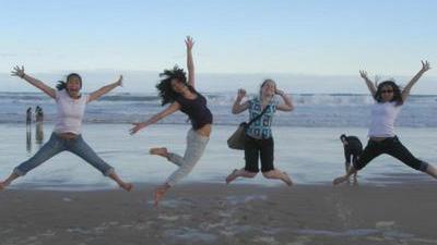 four young women leaping up in the air with their arms stretched out like stars, on a beach with glassy water in the background