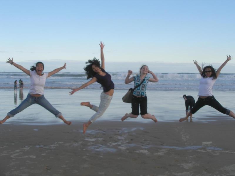 four young women leaping up in the air with their arms stretched out like stars, on a beach with glassy water in the background